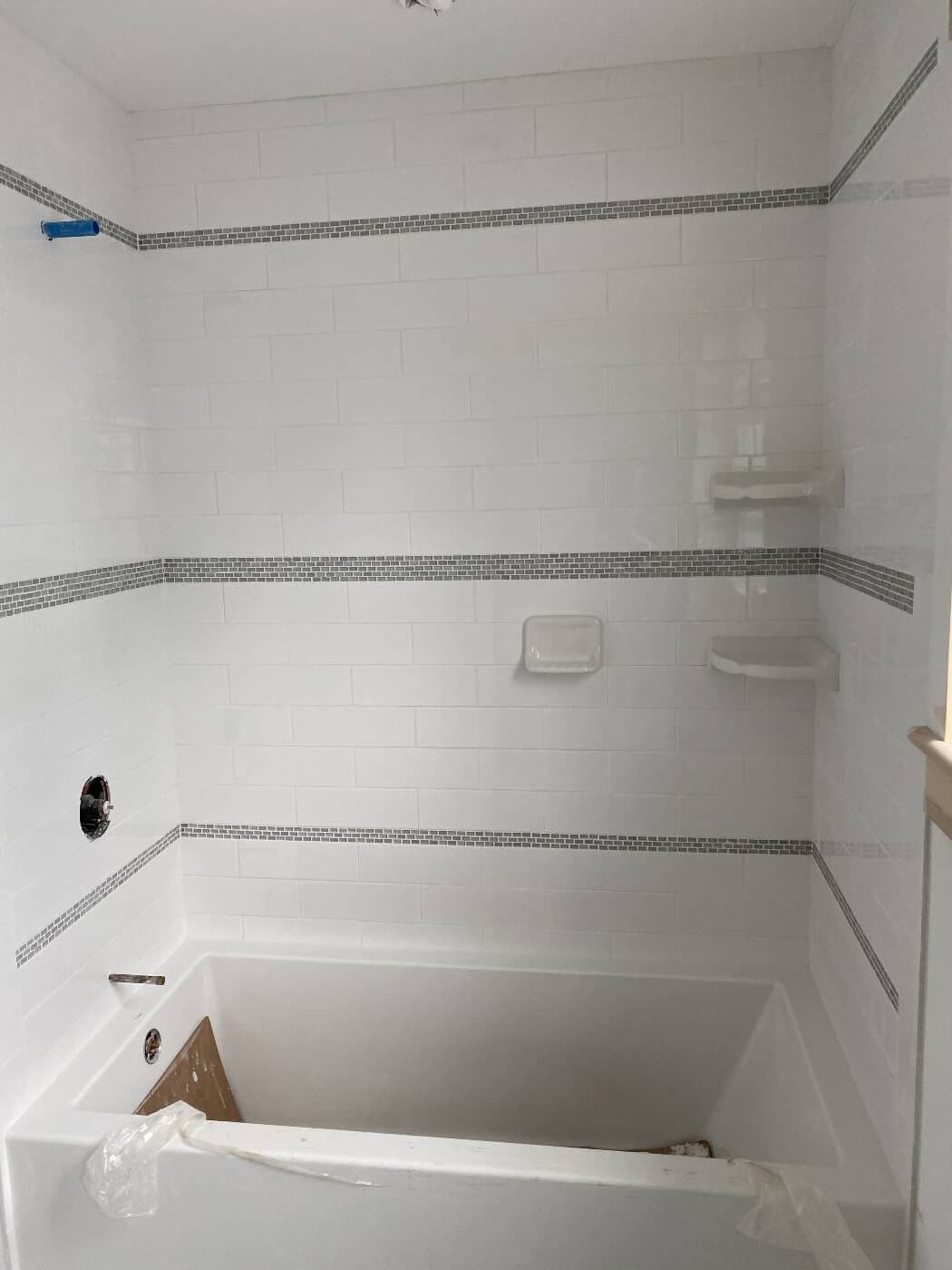 White tiled tub surround in progress with narrow mosaic bands, corner shelves and a recessed soap dish